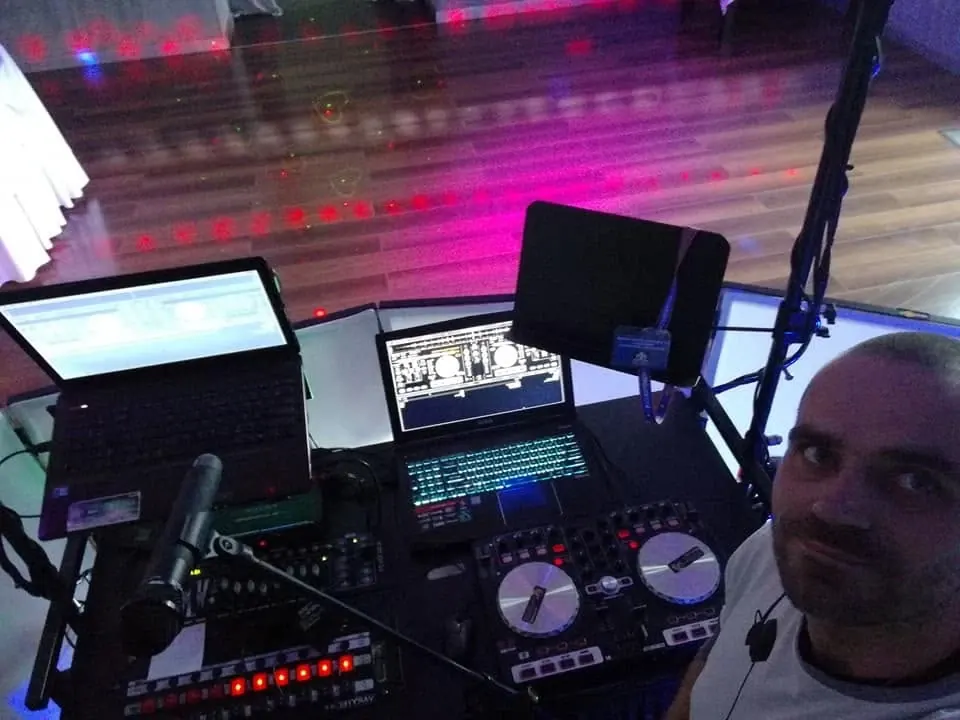 Stanowisko DJ Mastera na sali: dwa laptopy, kontroler i mikrofon, w tle parkiet w kolorowym świetle